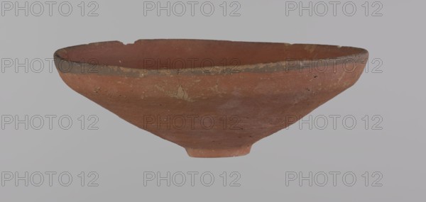 Bowl, Egyptian painted pottery bowl with conical shape and ring-shaped foot. Straight rim slightly flaring outward, irregular. Wheel-made from Nile clay type ND Vienna System. Surface even, glazed before firing; inner and outer rim have a narrow ~0.5 cm black band; remainder of vessel red. Traces of polish; glaze slightly worn in places—possible use-wear. Good condition with minor clay chips on rim and abrasions over surface. Analogies: Egypt's Golden Age no. 51, p. 78, ca. 1550–ca. 1425 BC; New Kingdom, early 18th Dynasty. Possible findplaces: Edfu, Deir el-Medina. Vessel height 5.3 cm; rim diameter 15.1 cm; bottom diameter 3.1 cm., A shallow, wide clay bowl with a rough texture resting against a pale gray background, showing a reddish-brown body with areas of darker brown along the rim and some lighter beige patches and speckles from wear; the bowl has an uneven, slightly chipped rim and a small raised base, with visible surface irregularities and subtle variations in the reddish, br...