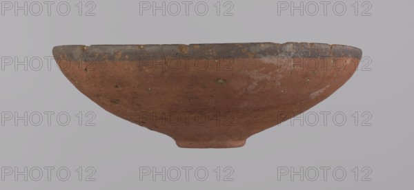 Bowl, A conical clay bowl with a ring-shaped bottom, wheel-made from Nile clay type NB2, Vienna System. The walls are even and covered with red engobe; the interior is polished. The rim is straight and slightly curled toward the center. A black painted band ~1 cm wide decorates the inside and outside of the rim. Vessel color: red. Preservation: quite good, with minor losses on the rim and small surface damages. Dimensions: height 5.5 cm; rim diameter 16.4 cm; bottom diameter 3.6 cm. Date: ca. 1550–1295 BC, New Kingdom, 18th Dynasty Tuthmosis III. Origin: Deir el-Medina. Type: ceramic vessel painted bowl., A shallow, rounded bowl with a wide rim shown in profile, exhibiting an earthy reddish-brown body with areas of darker brown and gray along the rim and scattered lighter spots and patches on the surface, a few small rough pits and irregularities in texture, and an overall matte, weathered appearance against a neutral gray background.