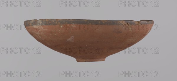 Bowl, A conical bowl with a ring-shaped ring bottom, wheel-made from Nile clay type NB2. Walls even, covered with red engobe; interior polished. Straight rim slightly curled toward the center; bottom wheel-modeled. Black painted bands ~1 cm wide on inside and outside of the rim. Vessel color red. Preservation: quite good, minor losses on the rim and minor surface damage. Comparative dating: New Kingdom, 18th Dynasty ca. 1550–1295 BC; Tuthmosis III. Dimensions: height 5.5 cm, rim diameter 16.4 cm, bottom diameter 3.6 cm., A shallow, rounded ceramic bowl with a worn rim showing a darker band along the edge and a predominantly reddish-brown body, displaying patches of lighter and darker discoloration and small specks, with areas of surface abrasion and rough texture, set against a neutral pale gray background; visible colors include reddish-brown, dark brown, pale gray, and small hints of white and greenish spots.