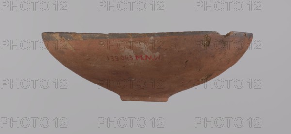 Bowl, A conical clay bowl with a ring-shaped bottom, wheel-made from Nile clay type NB2, Vienna System. Walls even, covered with red engobe; inner surface polished. Rim straight, slightly curled inward; bottom wheel-modeled. A black painted band ~1 cm wide on both the inside and outside of the rim. Vessel color red. Preservation: quite good, minor losses on the rim and minor surface damage. Comparative: New Kingdom, 18th Dynasty ca. 1550–1295 BC, Deir el-Medina; vessel height 5.5 cm, rim diameter 16.4 cm, bottom diameter 3.6 cm., A shallow, round ceramic bowl viewed from the side with an uneven rim and a small foot at the base; the bowl surface shows a worn, rough texture with chips and patches of abrasion. The visible colors are reddish-brown on the main body, darker brown and black along the rim, and small areas of pale off-white residue. The background is a smooth light gray.