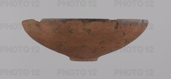 Bowl, Egyptian ceramic bowl with conical shape and ring-shaped bottom. Wheel-made from Nile clay type NB2 Vienna System. Surface even, covered with red engobe; inner surface polished. Straight rim slightly curled toward center; bottom wheel-modeled. Black painted band ~1 cm on inside and outside of rim. Vessel color red. Preservation: quite good, minor losses on rim and small surface damages. Comparative: New Kingdom, 18th Dynasty ca. 1550–1295 BC; Tuthmosis III, Deir el-Medina; vessel height 5.5 cm, rim diameter 16.4 cm, bottom diameter 3.6 cm., A shallow, rounded ceramic bowl with a wide rim and a small foot, shown against a pale gray background. The bowl's exterior surface is a warm reddish-brown with patches of darker brown and small areas of rough texture. The rim has a darker, almost blackened band along its top edge. The surface shows several irregular pits and spots of lighter tan and dark brown, giving it a weathered, earthy appearance. The background is a uniform light gray.