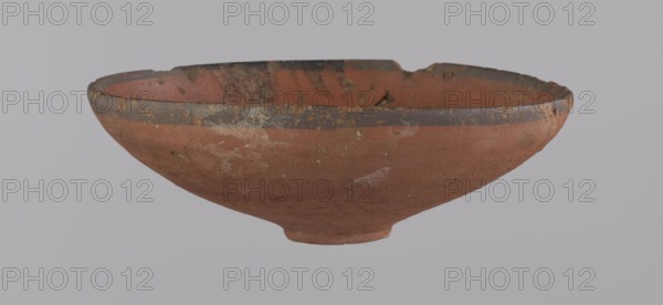 Bowl, A conical clay bowl with a ring-shaped ring bottom, wheel-made from Nile clay type NB2, Vienna System. The walls are even and covered with red engobe; the inner surface is polished. The rim is straight, slightly curled toward the center. A black painted band approx. 1 cm wide appears on the inside and outside of the rim. Vessel color: red. Preservation: quite good, minor losses on the rim and minor surface damages. Comparative dating: New Kingdom, 18th Dynasty ca. 1550–1295 BC; Tuthmosis III. Dimensions: height 5.5 cm; rim diameter 16.4 cm; bottom diameter 3.6 cm. Place of origin: Deir el-Medina, Egypt., A shallow, rounded ceramic bowl with a worn rim and uneven edge, showing surface chips and weathering; the bowl's exterior and interior display a terracotta color with patches of darker brown and areas of lighter, pale discoloration; the rim has a band of dark grayish-brown with small flecks of yellowish residue; the surface texture appears rough and pitted in places, with a smal...