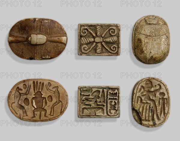 Amulet plaque with a rosette and the name Menkheperre. On one side: a sphinx on a gold sign, above it the symbol mn, and in front a cartouche of Menkheperre. On the other side: a rosette. Hieroglyphs/cryptograms; cartouches; rosette motif. Ca. 1550–1295 BC; New Kingdom, 18th Dynasty or later, Egypt. Plaque/amulet; height 1.9 cm, width 2.5 cm., inscriptions include the name Menkheperre., A neutral gray background holds a group of small carved objects arranged in a grid, each showing carved relief details. The top left object is an oval dark brown piece with a central raised elongated shape and textured carved lines radiating outward, showing lighter worn areas. The top center object is a small rectangular piece with a symmetrical carved motif of curved scrolls and ribbed elements in muted brown and beige tones. The top right object is an oval form resembling a scarab in mottled beige and light brown with subtle surface wear. The bottom left object is an oval light brown piece with a car...