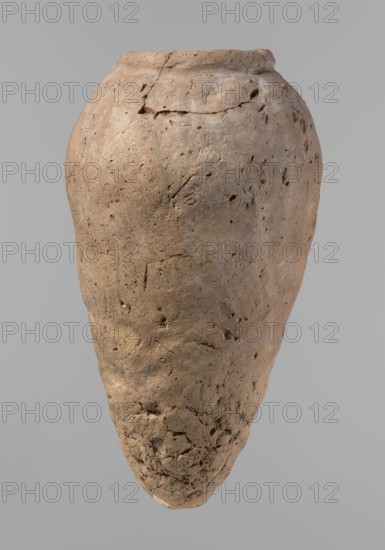 Vessel, Egyptian pottery, hand-made clay vessel, ceramics, Old Kingdom, c. 2686–c. 2181 BC, Edfu; vessel, height 31.9 cm, diameter 10.9 cm rim, diameter 17.4 cm body., A large, weathered pot with an elongated, tapered shape shown against a plain light gray background; the surface of the pot is rough and pitted with visible cracks and small holes, and its color ranges across shades of tan, beige, light brown, and muted gray with subtle darker brown spots and patches; the rim is slightly uneven and the texture near the base appears crumbly and more eroded compared with the smoother upper areas.