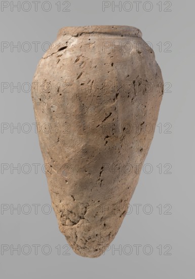 Vessel, Egyptian pottery, hand-made clay vessel, Old Kingdom c. 2686–c. 2181 BC, Edfu, vessel height 31.9 cm, rim diameter 10.9 cm, body diameter 17.4 cm., A single large clay vessel with a rough, pitted surface occupies the image, shown against a plain light gray background; the vessel has an elongated, tapered shape with a rounded shoulder and a slightly flaring rim, and its surface shows many small holes, cracks, and irregular textures. The visible colors are various shades of brown on the vessel, with subtle spots of darker brown and hints of gray, and the background is a uniform light gray.