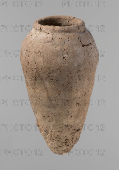 Vessel, Egyptian pottery, hand-made clay vessel, Old Kingdom c. 2686–c. 2181 BC, Edfu; comparative typology: Petrie Dendera pl. XVI.2; Hope Akhmim fig. 7 type AIVa 2bi; Reisner Mycerinus p. 212 fig. 64 3,4; Brunton Qau and Badari pl. LXXXI type 77N; Mace Naga ed-Der II figs. 87.11–14, 16–25; Reisner Naga ed-Der III fig. 28–33; Petrie Abydos II pl. XLIII, 79–80; Keiser, Avila, Dreyer, Jaritz, Laskowska-Kusztal, MDAIK 40 fig. 9b,c; vessel height 31.9 cm; rim diameter 10.9 cm; body diameter 17.4 cm., A weathered clay vessel with an elongated, tapered shape floating against a plain light gray background, showing an open rounded mouth with a thick rim and a rough, pitted surface; the vessel appears in shades of beige, tan, light brown, and small areas of darker brown, with visible cracks, small holes, and a more crumbly, textured base.