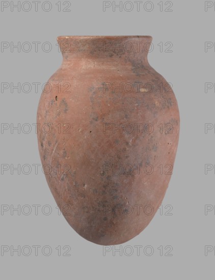 Vessel, Egyptian pottery vessel of clay, c. 2686–c. 2181 BC Old Kingdom?, from Edfu; height 16.5 cm, rim diameter 7.6 cm, belly diameter 11.2 cm., An oval-shaped clay vessel with a rounded body and a slightly flared neck, shown against a plain gray background; the surface shows a weathered texture with areas of worn and rough patches, small pits, and faint scratches; colors visible include reddish-brown as the dominant hue with patches of darker brown, grayish spots, and a few lighter beige flecks; overall the vessel appears matte and uneven in color with subtle mottling across its surface.