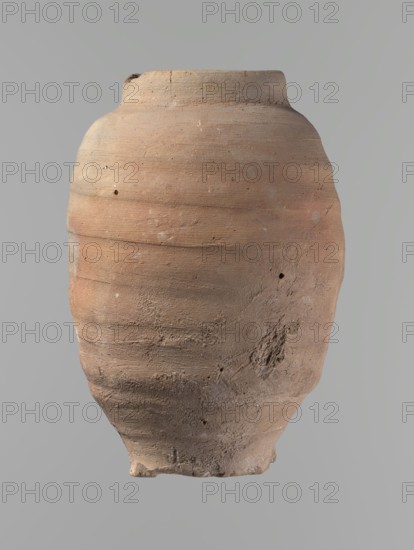 Vessel. Beer jug of a slightly irregular, conical shape with a wide, very short neck and a flat bottom. Flat, straight rim. Wheel-made from Nile clay of type NB2-ND Vienna System. The bottom is cut off with string, with clear potter's fingerprints around the bottom. Surface even. Clay color: red. Vessel complete, fairly well preserved, with minor damage to the rim and small chips on the surface. ca. 1550–ca. 1069 BC; New Kingdom, Edfu. Vessel. Height 24.4 cm; diameter rim 9.8 cm; diameter body 16.3 cm; diameter bottom 8.5 cm., A large, rounded clay vessel with a narrow neck and flared rim sits centered against a neutral gray background; the vessel surface shows an earthy palette of terracotta, pale brown, beige, and hints of dusty orange with areas of lighter and darker shading, visible horizontal banding and ridges from its construction, rough and pitted textures, several small holes and abrasions, and a few patches of uneven, crumbly surface near the lower half.