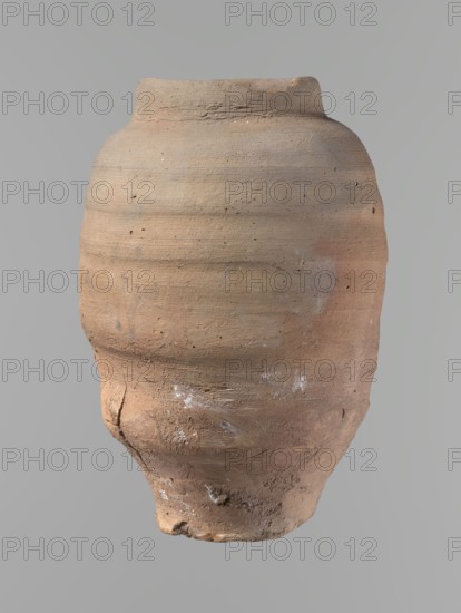 Vessel. Beer jug of slightly irregular, conical shape with a wide, very short neck and flat bottom. Flat, straight rim. Wheel-made from Nile clay of type NB2-ND Vienna System. Bottom cut off with string, with clear potter's fingerprints around the bottom. Surface even. Clay color: red. Vessel complete, fairly well preserved, with minor damage to the rim and small chips on the surface. Ca. 1550–1069 BC New Kingdom, Edfu. Vessel; height 24.4 cm; rim diameter 9.8 cm; body diameter 16.3 cm; bottom diameter 8.5 cm., An aged, rounded clay vessel occupies the image, showing a textured surface with visible horizontal striations and irregularities; its colors include shades of brown, tan, beige, and hints of gray with small patches of lighter white residue; the rim is slightly uneven and the body tapers toward a rough, eroded base with cracks and pitted areas, and the background is a flat, muted gray.