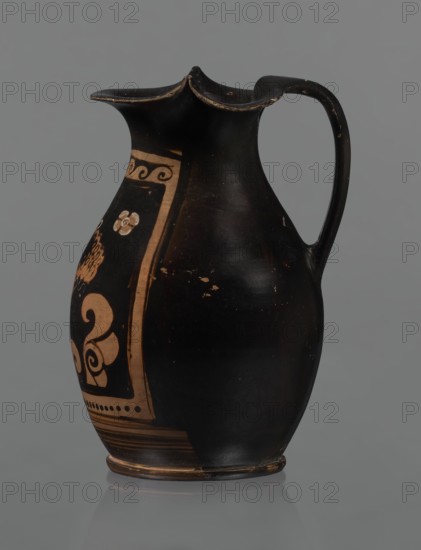 Red-figure oinochoe, Oinochoe with a flat foot separated by a ring, an oval belly, and sloping shoulders; wide, tripartite rim; smooth ribbon-like handle. Reddish clay with black glossy slip. Decoration: on the front of the belly, in a metopic field framed at the top with a breaking-wave ornament and at the bottom with a row of dots and lines — a woman's head in left profile wearing a cap sakkos with curly hair above the ears and a long bun of curls at the back tied with a taenia; the cap bears a diadem. Two half-palmettes flank the head both pointing same direction; on the right a spirally coiled tendril; two rosettes in the upper corners. Rosettes, necklace, taenia, earrings, and elements of the diadem painted in white. Good condition. End of the 4th century BC, Southern Italy. Oinochoe vessel, red-figure pottery, Greek., A dark ceramic jug with a curved handle and a flared, scalloped rim sits against a neutral gray background, the jug showing glossy black surfaces with areas of worn...
