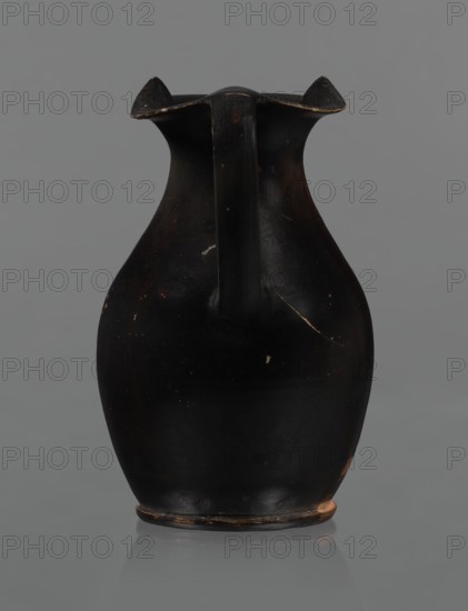 Red-figure oinochoe, Oinochoe with a flat foot separated by a ring, an oval belly, and sloping shoulders; wide, tripartite rim; smooth, ribbon-like handle. Reddish clay with black glossy varnish. Decoration: on the front of the belly, in a metopic field framed at top with a breaking-wave ornament and at bottom with a row of dots and lines, a woman's head in left profile wearing a cap sakkos with curly hair above the ears and a long bun of curls at the back tied with a taenia; a diadem on the cap. On either side of the head are two half-palmettes both pointing same direction; to the right a spirally coiled tendril; two rosettes in the upper corners. Rosettes, necklace, taenia, earrings, and elements of the diadem painted in white. Good condition. Late 4th century BC, Southern Italy., A dark, matte black ceramic jug with a rounded body, a narrow neck, and a single vertical handle running from the rim to the shoulder, showing small chips and worn edges where brownish-orange clay is expose...