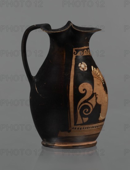 Red-figure oinochoe, Oinochoe with a flat foot separated by a ring, an oval belly, and sloping shoulders; wide tripartite rim; smooth ribbon-like handle. Reddish clay with black glossy varnish. Decoration: on the front of the belly, in a metopic field framed at top by a wave ornament and at bottom by a row of dots and lines — a woman's head in left profile wearing a cap sakkos with curly hair protruding above the ears and a long bun of curls at the back tied with a taenia; a diadem on the cap. On either side of the head are two half-palmettes both pointing same direction; on the right side a spirally coiled tendril; two rosettes in the upper corners. Rosettes, necklace, taenia, earrings, and elements of the diadem painted in white. Good condition. Late 4th century BC, Southern Italy., A black ceramic pitcher with a handle and a flared, scalloped rim standing against a gray background, the body showing a painted panel in a tan or light brown color with decorative swirls and the partial ...