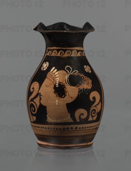 Red-figure oinochoe. Oinochoe with a flat foot separated by a ring, an oval belly, and sloping shoulders; wide tripartite rim; smooth, ribbon-like handle. Reddish clay with black glossy slip. Decoration: on the front of the belly, in a metopic field framed at the top by a breaking-wave pattern and at the bottom by a row of dots and lines, a woman's head in left profile wearing a cap sakkos with curly hair above the ears and a long bun of curls at the back tied with a taenia; a diadem on the cap. Two half-palmettes flank the head both pointing same direction; a spirally coiled tendril at the right side and rosettes in the upper corners. Rosettes, necklace, taenia, earrings, and parts of the diadem are painted in white. Good condition. Late 4th century BC, Southern Italy., A ceramic vessel with a flared, ruffled black rim and a rounded body sits against a gray background; the body shows a painted profile of a person in a terracotta color with black hair and ornamental black and terracott...