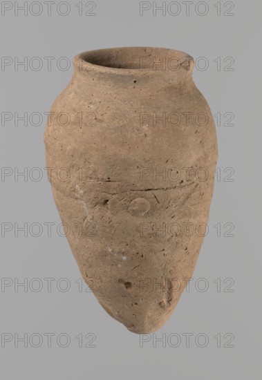 Vessel with hieratic inscription, Egyptian pottery conical clay vessel tapering toward the bottom, slightly irregular; widest at approx. 3/4 of the height. Narrow, sloping shoulders; short, wide neck; very gently rounded, widening rim; pointed bottom. Handcrafted; upper part shoulders, neck, rim turned. Around half the height a trace of joining of two separately made parts. Surface leveled, outer probably covered with a layer of engobe. Made of Nile clay Vienna System. Remains of a hieratic inscription in black on lower part of the belly. Rim damaged. Analogies cited; earlier dated to the 4th–5th Dynasty Old Kingdom, ca. 2686–2181 BC. Find from Edfu. Dimensions: height 13.2 cm; rim dia. 5.8 cm; body dia. 8.3 cm., A single weathered clay vessel with an elongated, tapered body and a rounded shoulder, shown against a plain gray background; the vessel surface is rough and pitted with small holes and irregularities, and its color appears brown with variations including lighter tan and some ...