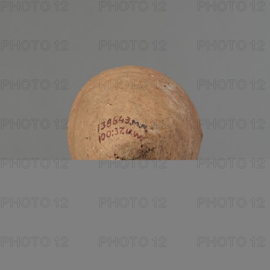 Vessel with hieratic inscription, Egyptian pottery ceramic conical vessel tapering towards the bottom, irregular, widest approx. 3/4 of the height. Narrow, slightly sloping shoulders; short, wide neck; very gently rounded, slightly flaring rim; pointed bottom. Shallow horizontal lines at the widest diameter made before firing. Handmade with upper part shoulders, neck, rim turned. Even surface, outer probably covered with engobe. Made of Nile clay NB2 Vienna System. Remains of a hieratic inscription in black on lower belly. Quite well preserved with slight damage to the rim. Analogies cited; earlier dated to the 4th–5th Dynasty. Old Kingdom, Edfu. Vessel height 13.9 cm; rim diameter 5.9 cm; belly diameter 8.5 cm., A rounded earthen object with a rough, textured surface in shades of brown and tan sits against a flat gray background; the object shows small areas of darker brown and faint pale spots, with a few small pits and irregularities on its surface.