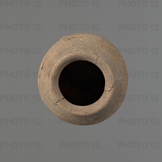 Vessel with hieratic inscription, Egyptian pottery, ceramics, clay conical vessel tapering towards the bottom, irregular, widest approx. 3/4 of the height. Narrow, slightly sloping shoulders; short, wide neck; very gently rounded, slightly flaring rim; pointed bottom. Shallow horizontal lines at the widest diameter made before firing. Handmade vessel; upper part shoulders, neck, rim turned. Even surface, outer probably covered with a layer of engobe. Made of Nile clay NB2 Vienna System. Remains of a hieratic inscription in black on lower part of the belly. The vessel is glued from original fragments. Old Kingdom, Edfu. Height 14.8 cm; rim diameter 6.2 cm; belly diameter 9.1 cm. Comparable types dated ca. 2686–ca. 2181 BC; earlier parallels assigned to 4th–5th Dynasty., A round, weathered clay vessel viewed from above with a wide dark opening at its center, showing a rough, cracked surface and uneven rim; the vessel is primarily brown with variations of tan and gray, and it sits against...