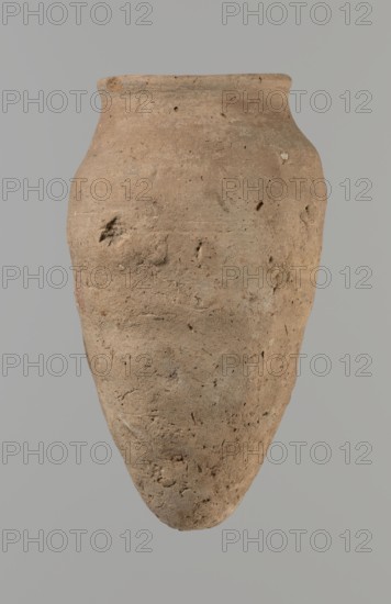 Vessel with hieratic inscription. Conical Nile-clay vessel NB2, Vienna System, handcrafted with upper part turned; tapering to a pointed bottom, irregular, widest at ~3/4 height. Narrow, slightly sloping shoulders; short, wide neck; very gently rounded, slightly flaring rim. Smooth surface, externally probably covered with engobe. Remains of a hieratic inscription in black on lower belly. Fair condition with slight damage to rim. Old Kingdom, ca. 2686–2181 BC. Height 13.9 cm; lip diameter 5.9 cm; body diameter 8 cm. Analogies: parallels in Petrie Dendera, Abydos, Hope Akhmim, Reisner Mycerinus; Naga ed-Der, Brunton Qau and Badari, Mace Naga ed-Der II, Keiser et al., An elongated, rounded clay vessel with a narrow neck and flared rim fills the image, showing a rough, pitted surface with irregular indentations and small embedded fragments; the vessel is primarily light brown and tan with subtle variations of beige and darker brown speckles, photographed against a smooth, neutral gray bac...