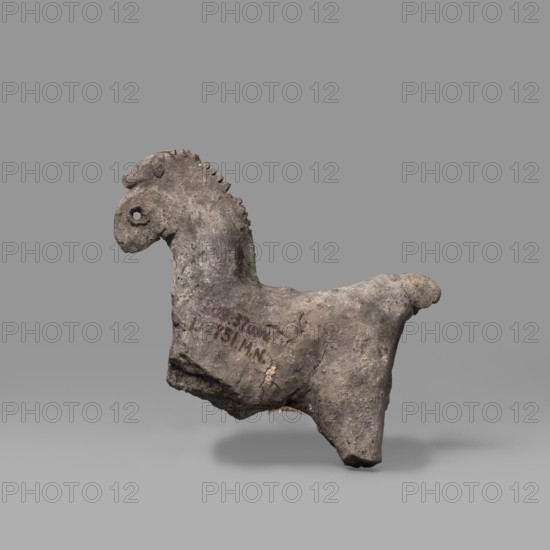 Horse figurine, Schematic representation of a horse with a short torso, a massive arched neck and a crest-like mane. Small head with eyes marked by drilled holes. Raised tail preserved in the upper part. Dated 664–30 BC Late Period–Ptolemaic Period. From Edfu. Terracotta figurine, height 8 cm, width 9 cm., A roughly shaped, weathered object resembling an animal form with a curved neck and head, a short mane of jagged ridges along the neck, a round eye hole, and a stubby tail; the surface is textured and uneven with visible chips and worn areas, showing a mix of gray and brown tones with darker and lighter patches; it rests against a uniform medium-gray background and casts a soft shadow beneath it.