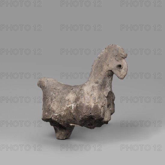 Horse figurine. Schematic representation of a horse with a short torso, a massive arched neck and a crest-like mane; small head with eyes marked by a drilled hole; raised tail preserved in the upper part. Late Period–Ptolemaic Period, 664–30 BC. From Edfu. Terracotta figurine, height 8 cm, width 9 cm., A small sculptural object resembling an animal form sits against a smooth gray background; the piece appears rough and irregular with a textured surface showing wear and pitting, and its overall color palette includes shades of gray, brown, and muted beige. The object has a raised head with a rounded snout and a hollowed area where an eye might be, a short neck with a scalloped ridge along the top, a broad body that tapers toward a stubby tail, and short, thick legs providing support. Surface variations show lighter and darker patches, small pits, and areas of rougher, crumbly texture, with some crusty deposits and subtle scratches visible on the body.