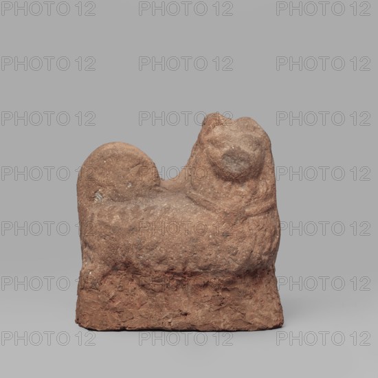 Dog figurine, dogs, 1st–2nd century Roman period, from Edfu; terracotta figurine, height 6.6 cm, width 6.5 cm., A small rough-textured sculpture of a reclining animal-like form sits against a plain background; the object has an overall reddish-brown hue with variations of lighter tan and occasional pale flecks, a rounded head with a subtle snout and ear-like protrusions, a curved back rising into a rounded hump, and a blocky rectangular base with an irregular, crumbly surface.