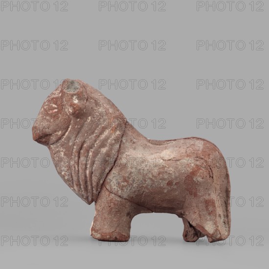 Bull figurine, Roman period, Tell Atrib, figurine, height 12.5 cm, width 16 cm, A sculpted animal figure standing on a flat surface, shown in profile facing left, with a rounded body, short sturdy legs, and a pronounced mane that drapes along the neck; the surface is rough and weathered with visible cracks and chips, displaying a palette of reddish-brown, pinkish, beige, and patches of gray, with areas of lighter and darker wear that create mottled texture.