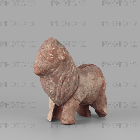 Bull figurine, Roman period, Tell Atrib, figurine, height 12.5 cm, width 16 cm, A small weathered sculpture of an animal standing on a flat surface, rendered in a rough reddish-brown material with areas of lighter beige and pale gray, showing a rounded head with a simplified face and a ridged mane or textured neck, a compact body with visible surface wear and chips, stout legs with one front leg showing a lighter interior, and a smooth neutral gray background.