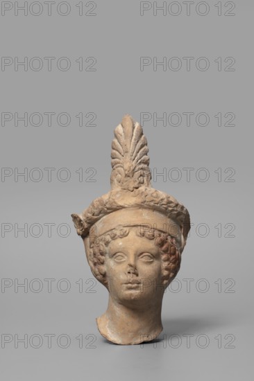 Head of a Dionysus figurine; maker, Tarentine workshop; Dionysus mythology, wreaths; Greek deity; 5th–4th century BC previously 2nd half of the 5th century BC; Tarentum, Southern Italy Italy; sculpture; height 12.9 cm, width 7 cm, depth 6 cm, weight 0.18 kg., A terracotta head of a person with a tall decorative headdress shaped like a stylized plume sits against a neutral gray background; the head is light brown with subtle darker brown and reddish patches, showing detailed curly hair framing the face, closed or slightly open eyes, a defined nose and lips, and a neck base that appears broken or incomplete, while the headdress has textured leafy or feathered elements and a band encircling the forehead.
