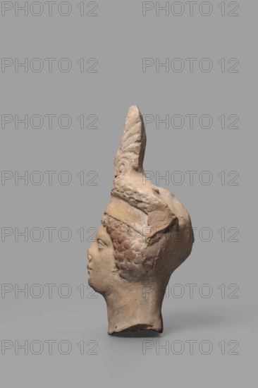 Head of a Dionysus figurine, unknown creator; unknown Tarentine workshop; Dionysus mythology, wreaths, Greek deities; 5th–4th century BC previously dated to 2nd half of the 5th century BC; Tarentum, Southern Italy Italy; sculpture; height 12.9 cm, width 7 cm, depth 6 cm, weight 0.18 kg., A profile view of a sculpted head in a beige to light brown clay tone against a solid gray background, showing textured curly hair, a smooth forehead and nose, full lips, and a defined chin and neck; the head wears a tall, pointed headdress with visible carved ridges and ornamental texture, and there are areas of darker brown and subtle reddish-brown discoloration on the hair and headdress as well as lighter patches of pale beige on the face and neck.