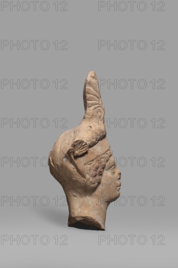 Head of a Dionysus figurine, unknown Tarentine workshop, Dionysus mythology, wreaths, Greek deities, 5th–4th century BC previously dated to 2nd half of the 5th century BC, Tarentum, Southern Italy Italy; sculpture; height 12.9 cm, width 7 cm, depth 6 cm, weight 0.18 kg., A sculpted head in a reddish-brown terracotta tone is shown in profile against a smooth gray background; the figure wears a tall, pointed helmet with a decorative crest and a small fan-shaped ornament at the temple, and the face displays a calm expression with a defined nose, lips, and curly hair around the forehead, while the neck and lower edge of the bust show rough, broken edges and the surface has patches of lighter and darker wear in shades of beige and pale gray.