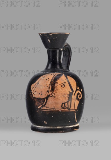 Aryballus lekythos with a depiction of a woman's head in a cap, ca. 400 BC, Attica, vessel lekythos, height 8.8 cm, width 5.3 cm, A small rounded ceramic vessel with a narrow neck, flared rim, and single curved handle sits against a plain background; the vessel is primarily black with areas of warm terracotta where the surface decoration shows a profile of a human face wearing a head covering and an earring, with hair detail and ornamental curling lines beside the face; the black surface has small chips and worn spots revealing the terracotta beneath, and the base shows similar wear, with the overall colors being black and terracotta against a gray backdrop.