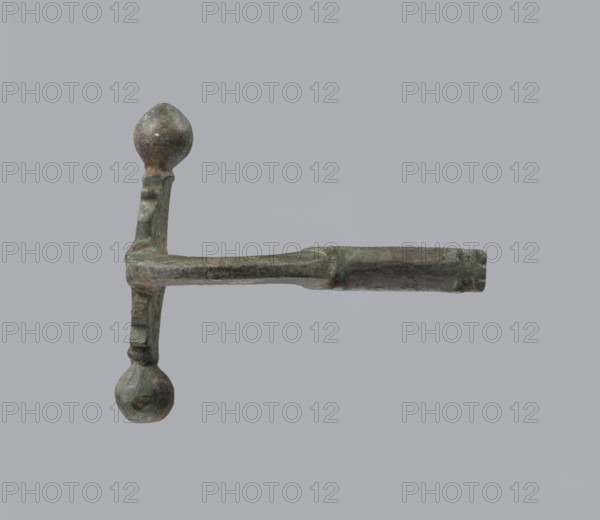 Onion-shaped fibula, Italy. Bronze fibula with a hinged structure featuring an arched ring made from a quadrilateral plate, terminated on one side by a thick transverse plate with a stepped upper edge and large balls on both sides, and on the other by a thick sheath formed as a half-cylinder, cut to insert a needle. Condition: good; surface covered with greenish corrosion and brown spots. Date: late 3rd–4th century; probable origin: Rome. Jewelry, fibula., A single metallic object with a horizontal shaft and a perpendicular shorter arm forming a T-like shape, each end of the shorter arm terminating in a rounded knob; the object has a rough, pitted surface and an uneven patina showing shades of green and brown against a uniform light gray background.