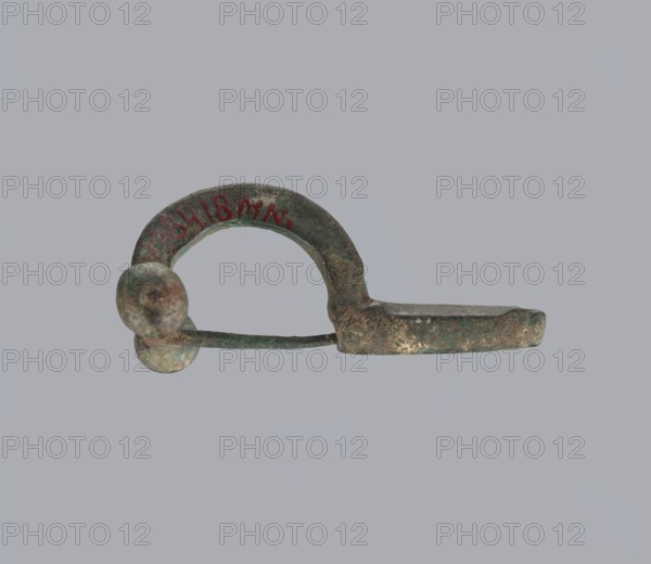 Onion-shaped fibula, Italy; bronze. A fibula with a hinged structure featuring an arched ring made from a quadrilateral plate, terminated on one side by a thick transverse plate with a stepped upper edge and large balls on both sides, and on the other by a thick sheath formed as a half-cylinder, cut to receive a needle. Condition: good; surface covered with greenish corrosion with brown spots. Date: late 3rd–4th century; origin possibly Rome. Jewelry, fibula., An aged metal object with a curved arch and an attached straight bar, showing a weathered, pitted surface and greenish patina, with two rounded knob-like ends on one side; colors visible include green, brown, gray, and hints of reddish discoloration, set against a uniform light gray background.