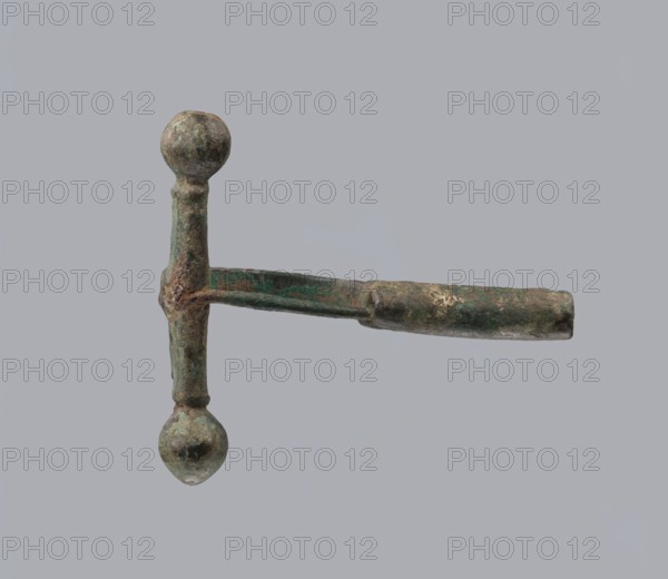 Onion-shaped fibula, Italy. Bronze fibula with hinged structure: an arched ring made from a quadrilateral plate, one end terminating in a thick transverse plate with a stepped upper edge and large balls on both sides, the other end a thick sheath shaped as a half-cylinder, cut to insert a needle. Condition: good; surface covered with greenish corrosion and brown spots. Late 3rd–4th century. Jewelry, fibula., An object with a vertical shaft and a horizontal arm forming a T-shape, showing rounded knobs at the top and bottom of the shaft and a tubular extension on the horizontal arm; the surface appears rough and corroded with mottled green and brown patina and some darker and lighter patches, set against a uniform light gray background.