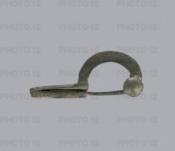 Onion-shaped fibula, Italy. Bronze fibula with a hinged structure and an arched ring made from a quadrilateral plate; one end terminates in a thick transverse plate with a stepped upper edge and large balls on both sides, the other end has a thick sheath formed as a half-cylinder, cut to insert a needle. Condition: good, surface with greenish corrosion and brown spots. Late 3rd–4th century, Rome . Jewelry, fibula., A single small metal object with a curved arch and a rounded knob at one end sits against a plain gray background; the object shows a patina with shades of green, brown, and dark gray, with some areas appearing mottled and worn, a thin flat flange extends from beneath the arch and a narrow straight bar connects the arch to the knob, the surfaces show irregular texture and some rough edges.