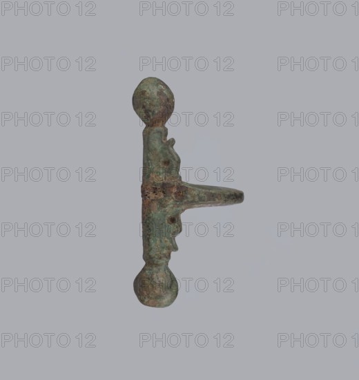 Onion-shaped fibula, Italy. A fibula with a hinged structure featuring an arched ring formed from a quadrilateral plate, ending on one side with a thick transverse plate with a stepped upper edge and large balls on both sides, and on the other side with a thick sheath shaped as a half-cylinder, cut to accept a needle. Condition: good; surface covered with greenish corrosion and brown spots. Late 3rd–4th century, possibly Rome. Jewelry, bronze fibula., An elongated metal object with a weathered, rough surface and a greenish patina, showing areas of brown and darker green; it has bulbous rounded ends, a central shaft with irregular protrusions and holes, and a flat horizontal extension projecting from one side; the background is a uniform pale gray.