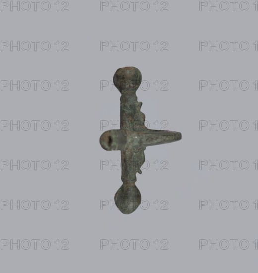 Onion-shaped fibula, Italy; bronze fibula with hinged structure: an arched ring formed from a quadrilateral plate, one end terminating in a thick transverse plate with a stepped upper edge and large balls on both sides, the other ending in a thick sheath shaped as a half-cylinder, cut to receive a needle. Condition: good, surface with greenish corrosion and brown spots. Date: late 3rd–4th century; probable origin: Rome. Jewelry, fibula., A single metal object is centered against a plain light gray background, showing a vertical shaft with rounded knobs at both ends and a horizontal protruding arm; the object has a weathered, mottled surface with greenish patina and areas of brown and dark gray, and irregular textured edges and pitted spots are visible along its form.