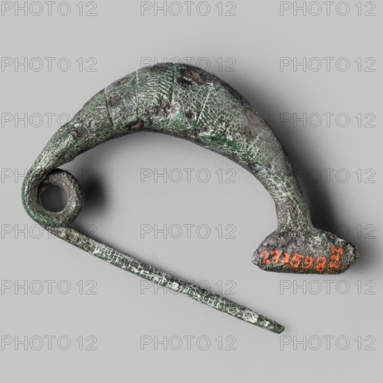 Leech fibula with a short stem, Bronze fibula with an arched thickening and a full bow. Short clasp foot made of an oval plate which, when bent, forms a hook for a short pin. The bow is decorated with a series of transverse parallel engraved lines forming stripes alternately smooth and diagonally hatched with engraved lines. Dated to the 8th–7th century BC 2nd half of the 8th–early 7th century BC, Etruria. Fibula, height 4.4 cm., An aged metallic object with a curved, hook-like main piece and a separate straight pin-like element lying across it, both showing a rough textured surface with visible corrosion and a mottled green patina, along with areas of dark brown and gray; the background is a smooth light gray.