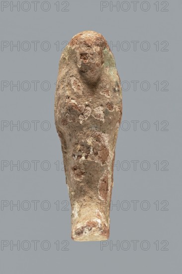 Ushabti. A mummy-shaped ushabti figurine made of clay, impressed in a mold. Traces of white primer and paint in places on the surface. The back of the figurine is unfinished. The arms are separated, the hands are crossed on the chest, probably the left folded over the right. In both hands the figurine holds hoes — the right much more clearly visible, the left very indistinct. The tools are painted black. A three-part wig on the head, the strand at the back is not separated. The ears are invisible. Facial details illegible. Inscription illegible/missing. Figurine not very well preserved — significant paint loss over the entire surface. Typology according to Schneider: 4.3.2 VIIIA2/W.16 H.29 I.5 B.0 Tp.0. ca. 1069–715 BC; Third Intermediate Period, 21st–23rd Dynasty ca. 1069–715 BC, Egypt. Shabti. Height 9.4 cm; width 3.1 cm; depth 2.7 cm., A weathered vertical sculpture of a simplified human figure with a rounded head and indistinct facial features, displaying a rough, pitted surface an...
