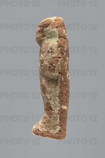Ushabti. A mummy-shaped ushabti figurine made of clay, impressed in a mold. Traces of white primer and paint remain in places on the surface. The back of the figurine is unfinished. The arms are separated, hands crossed on the chest, probably left folded over right. In both hands the figurine holds hoes — the right one much clearer, the left very indistinct; the tools are painted black. A three-part wig on the head, the strand at the back is not separated. Ears invisible; facial details illegible. Inscription illegible/missing. Figurine not very well preserved — significant paint loss over the entire surface. Typology according to Schneider: 4.3.2 VIIIA2/W.16 H.29 I.5 B.0 Tp.0. Date: ca. 1069–715 BC, Third Intermediate Period, 21st–23rd Dynasty XXI–XXV Dynasty, Egypt. Type: shabti. Dimensions: height 9.4 cm, width 3.1 cm, depth 2.7 cm., A vertical, elongated rough object with an irregular rounded top and a slightly flared base set against a flat gray background; the surface shows a mix...
