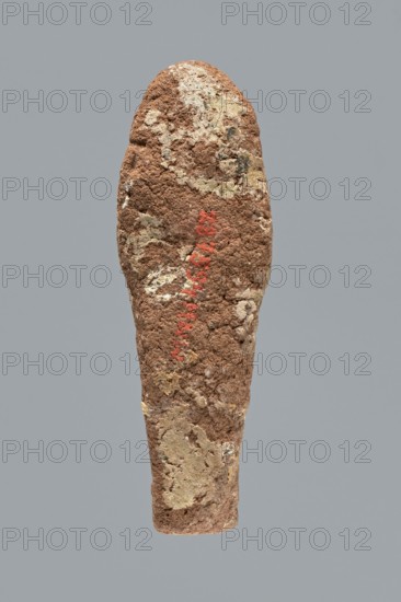 Ushabti. A mummy-shaped ushabti figurine made of clay, impressed in a mold. Traces of white primer and paint remain in places; significant overall paint loss. The back is unfinished. Arms are separate, hands crossed on the chest with the left likely folded over the right. The figurine holds hoes in both hands — the right hoe is much clearer, the left indistinct; the tools are painted black. Head bears a three-part wig with the rear strand not separated; ears invisible; facial details illegible. Inscription illegible/missing. Typology Schneider: 4.3.2 VIIIA2/W.16 H.29 I.5 B.0 Tp.0. Date ca. 1069–715 BC, Third Intermediate Period 21st–23rd Dynasty. Type: shabti. Dimensions: height 9.4 cm, width 3.1 cm, depth 2.7 cm., A roughly elongated, tapering object with a textured, crumbly surface in shades of brown and tan, with patches of lighter beige and pale yellow; there is a vertical area of muted red pigment on the surface; the background is a flat, uniform gray.