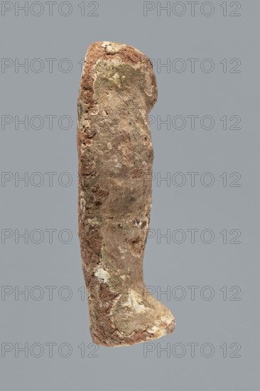Ushabti. Mummy-shaped ushabti figurine made of clay, impressed in a mold. Traces of white primer and paint in places on the surface. The back of the figurine is unfinished. Arms are separated; hands crossed on the chest, probably left folded over right. Figurine holds hoes in both hands — the right much more clearly visible, the left indistinct. Tools painted black. Three-part wig on the head, the strand at the back is not separated. Ears not visible. Facial details illegible. Inscription illegible/missing. Figurine not very well preserved — significant paint loss over the entire surface. Typology according to Schneider: 4.3.2 VIIIA2/W.16 H.29 I.5 B.0 Tp.0. ca. 1069–715 BC; Third Intermediate Period, 21st–23rd Dynasty ca. 1069–ca. 715 BC, Egypt. Shabti. Height 9.4 cm; width 3.1 cm; depth 2.7 cm., A single upright weathered object with an irregular elongated shape, showing a rough textured surface with patches of brown, tan, beige, off-white, and small areas of muted green, photographed...