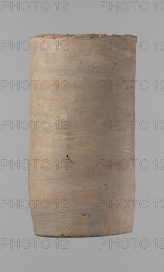 Canopic urn. Clay canopic urn with a cylindrical shape and a flat bottom and a flat, straight rim. Made on the wheel of Nile clay type NB2 Vienna System. Bottom is cut off with a knife; surface leveled. On the outer wall is a rectangle painted in red, measuring 11.5 cm width by 11 cm height. Color of vessel: light brown. Preservation condition quite good — rim partially filled with plaster and painted. Date ca. 1550–1069 BC New Kingdom, provenance Deir el-Medina. Vessel; height 23.5 cm, diameter 12.5 cm rim, diameter 11.4 cm bottom., An upright cylindrical object with a slightly uneven rim and base, showing a rough, textured surface with small pits and scratches; the object displays a range of muted earth tones including beige, tan, light brown, and subtle hints of gray and pale orange, with faint irregular darker spots and thin hairline cracks visible across its surface.