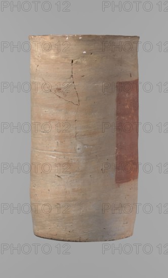 Canopic urn, Clay canopic urn with a cylindrical shape and a flat bottom and a flat, straight rim. Vessel made on the wheel of Nile clay type NB2 Vienna System. Bottom is cut off with a knife; surface leveled. On the outer wall is a rectangle painted in red, measuring 11.5 cm width by 11 cm height. Color of vessel: light brown. Preservation quite good — rim partially filled with plaster and painted. Date: ca. 1550–ca. 1069 BC New Kingdom, provenance: Deir el-Medina. Function: canopic urn. Dimensions: height 23.5 cm; diameter 12.5 cm rim; diameter 11.4 cm bottom., A tall cylindrical ceramic vessel with a matte, weathered surface showing a range of earthy colors including beige, light tan, pale cream, and a vertical patch of muted reddish-brown on one side; the surface has visible cracks, small pits, and irregular texture variations, with subtle horizontal striations and areas of slightly darker and lighter tones, and a top rim that is uneven and worn.