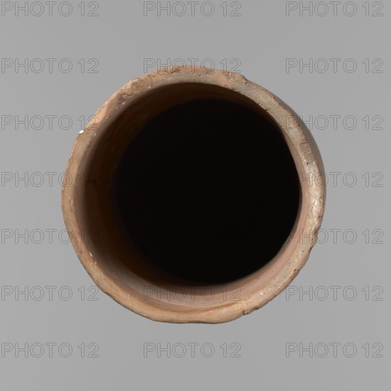 Canopic urn. Clay canopic urn with a cylindrical shape and a flat bottom and a flat, straight rim. Vessel made on the wheel of Nile clay type NB2 Vienna System. The bottom is cut off with a knife; surface leveled. On the outer wall is a rectangle painted in red, measuring 11.5 cm width by 11 cm height. Color of vessel: light brown. Preservation condition: quite good — rim partially filled with plaster and painted. Date: ca. 1550–ca. 1069 BC New Kingdom. Place of origin: Deir el-Medina. Function/type: canopic vessel. Height 23.5 cm; diameter rim 12.5 cm; diameter bottom 11.4 cm. Maker: unknown., A top-down view of a round, open clay vessel with an uneven rim and rough texture, showing colors of brown, tan, beige, gray, black, and small patches of white; the interior appears very dark to black while the background is a uniform light gray.