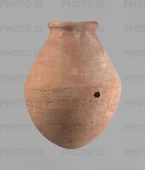 Vessel, Egyptian ceramic vessel with an ovoid belly, a wide short neck and a rounded bottom; straight, upturned rim. Wheel-made from Nile clay of type NB2 Vienna System. Surface levelled and outer surface covered with engobe. Outer surface red, inner surface yellowish-brown. Firing hole at widest circumference approx. 1 cm diameter. Well-preserved. Date approx. 1550–1295 BC; New Kingdom, 18th Dynasty. From Deir el-Medina. Vessel height 27 cm; rim diameter 11 cm; body diameter 20.5 cm., A rounded clay vessel with a narrow neck and flared rim fills the image, showing a textured surface with visible scratches and imperfections; the vessel is primarily terracotta and earthy orange with areas of lighter beige and dusty white, and there is a small irregular hole on one side; the background is a uniform light gray.