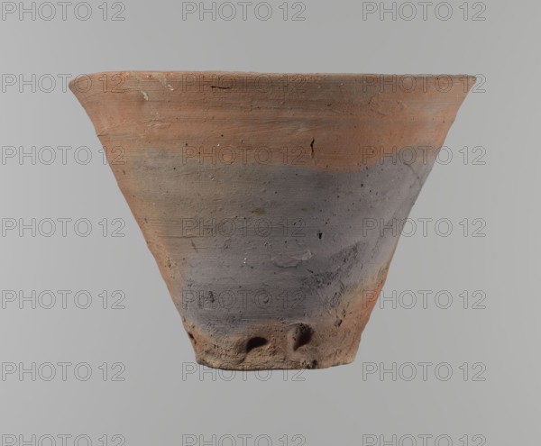 Bowl. Vessel flower pot with a conical shape and a flat bottom. Irregular walls. Straight, flat, slightly flaring, asymmetrical rim. Made on the wheel of Nile clay type NB2 Vienna System. Irregular bottom, cut off with a string; a 3.2 cm diameter hole in the bottom. Potter's fingerprints present in the lower part near the bottom. Smooth surface. Reddish-brown clay. Condition good, with only minor damage to the rim. Ca. 1550–1069 BC New Kingdom, Deir el-Medina. Height 18 cm; diameter 15.8 cm rim, 9.5 cm bottom., A clay vessel with a flared rim and conical shape is shown against a plain background; its surface has a rough, textured appearance with areas of terracotta orange and muted gray, and there are a few irregular pits and small holes near the base as well as some scratches and surface imperfections.