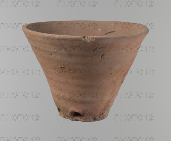 Bowl. Vessel flower pot with a conical shape and a flat bottom. Irregular walls. Straight, flat, slightly flaring, asymmetrical rim. Made on the wheel of Nile clay type NB2 Vienna System. Irregular bottom, cut off with a string; 3.2 cm diameter hole in the bottom. Clear potter's fingerprints in the lower part next to the bottom. Smooth surface. Reddish-brown color. Condition good, only minor damage to the rim. Ca. 1550–1069 BC, New Kingdom, Deir el-Medina. Height 18 cm; rim diameter 15.8 cm; bottom diameter 9.5 cm., A terracotta-colored clay pot with a conical shape, showing a rough textured surface with small pits, scratches, and irregularities; the rim is slightly uneven and has a few chips, and the base appears worn with an indentation and rough edges; colors visible include terracotta, brown, reddish-brown, and small areas of gray and lighter beige, all set against a neutral light gray background.