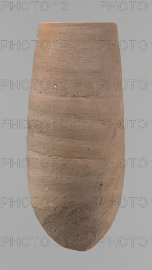 Vessel with a conical shape and a pointed bottom. Straight, tapering rim. Made on the wheel of Nile clay type NB2 according to the Vienna System classification. Bottom and outer surface worked with a hard tool. Surface even. Light brown color. Fairly well preserved, with minor damage on the rim. c. 1550–c. 1069 BC, New Kingdom, Deir el-Medina. Vessel; height 20.8 cm, diameter 7 cm lip, 9 cm body., A tall, elongated clay object with a roughly cylindrical, slightly tapered form shown against a plain background, exhibiting a textured, weathered surface with visible horizontal tool marks and small pits. The surface shows a range of earthy tones including light brown, tan, beige, and hints of gray, with some lighter specks scattered across it. The top edge appears flat and the bottom narrows to a rounded point, with variations in surface roughness and subtle color shifts along the object's vertical axis.