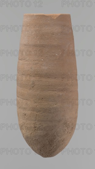 Vessel with a conical shape and a pointed bottom. Straight, tapering rim. Made on the wheel of Nile clay type NB2 Vienna System classification. Bottom and outer surface worked with a hard tool; surface even. Light brown color. Fairly well preserved, minor damage on the rim. c. 1550–c. 1069 BC, New Kingdom, Deir el-Medina. Vessel, height 20.8 cm, diameter 7 cm lip, diameter 9 cm body., A single tall, rounded clay vessel occupies the image, shown from top to bottom against a neutral gray background; the vessel has a rough, textured surface with visible horizontal striations and small pits, an uneven rim with a small chipped area, and an overall matte finish; the dominant color of the vessel is earthy brown with subtle variations of lighter and darker brown and occasional tiny pale flecks, while the background is a flat gray.