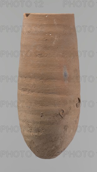 Vessel, Vessel with a conical shape and a pointed bottom; straight, tapering rim. Wheel-made of Nile clay type NB2 Vienna System. Bottom and outer surface worked with a hard tool; surface even. Light brown color. Fairly well preserved, minor damage on the rim. c. 1550–c. 1069 BC; New Kingdom, Deir el-Medina. Vessel; height 20.8 cm; diameter 7 cm lip, 9 cm body., An elongated oval clay vessel with a rough, textured surface, showing a few small chips and indentations around its rim and body; the vessel appears predominantly terracotta brown with subtle variations including patches of darker brown and a muted grayish-blue smudge on one side, plus scattered tiny white flecks; the background is a flat light gray.